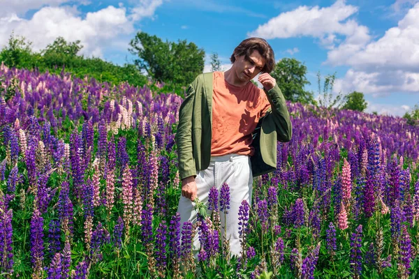 Tall handsome man in a green jacket standing on lupine flowers field holding lupine flowers in hand, enjoing the beauty of nature. Man surrounded by purple and pink lupines.