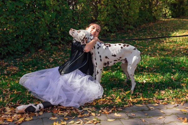 Ballerina with Dalmatian dog in the golden autumn park. Woman ballerina in a white ballet skirt and black leather jacket and in pointe shoes in autumn park hugging her spotty dalmatian dog.