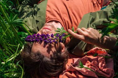 Yeşil ceketli uzun boylu yakışıklı adam lupine çiçeklerinin arasında yatıyor. Üst görünüm.