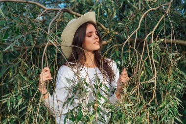 Beyaz kazak, kot pantolon ve kovboy hasır şapkası giymiş genç, güzel esmer kadın yeşil yapraklı ağaç brunch 'larına yakın duruyor.