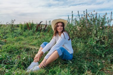 Beyaz kazak, kot pantolon ve kovboy hasır şapkası giyen genç, güzel esmer kadın yeşil çimlerin üzerinde oturuyor, gün batımında gülümsüyor ve gülüyor.