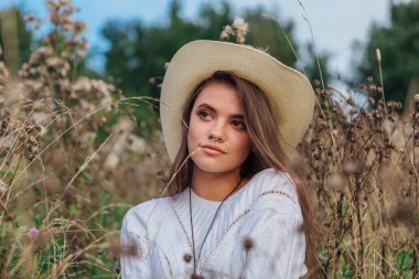 Beyaz kazak, kot pantolon ve kovboy hasır şapkası giyen genç, güzel esmer kadın kuru kahverengi çimlerin üzerinde oturuyor, gün batımında gülümsüyor ve gülüyor.