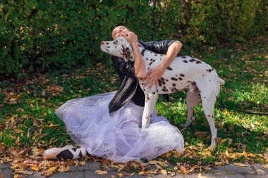 Altın sonbahar parkında Dalmaçyalı bir köpekle balerin. Beyaz balerin etekli, siyah deri ceketli, sonbahar parkında sivri uçlu ayakkabılı, benekli dalmaçyalı köpeğine sarılan bir kadın balerin..