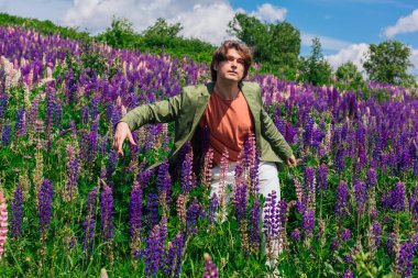 Yeşil ceketli uzun boylu yakışıklı bir adam lupin çiçeği tarlasında dans ediyor, doğanın güzelliğini emrediyor. Mor ve pembe lupinlerle çevrili bir adam..