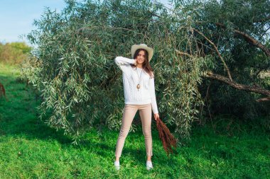 Beyaz kazak, kot pantolon ve kovboy hasır şapkası giymiş genç, güzel esmer kadın elinde kuru yapraklarla ağaç brunch 'larına yakın duruyor.