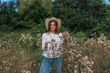 Beyaz kazak, kot pantolon ve kovboy hasır şapkası giymiş genç, güzel esmer kadın kuru kahverengi çimlerin üzerinde duruyor, gün batımında gülümsüyor ve gülüyor.