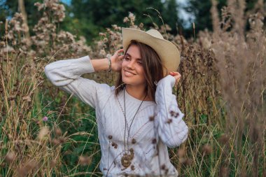 Beyaz kazak, kot pantolon ve kovboy hasır şapkası giyen genç, güzel esmer kadın kuru kahverengi çimlerin üzerinde oturuyor, gün batımında gülümsüyor ve gülüyor.