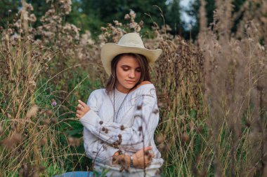 Beyaz kazak, kot pantolon ve kovboy hasır şapkası giyen genç, güzel esmer kadın kuru kahverengi çimlerin üzerinde oturuyor, gün batımında gülümsüyor ve gülüyor.