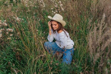Beyaz kazak, kot pantolon ve kovboy hasır şapkası giyen genç, güzel esmer kadın kuru kahverengi çimlerin üzerinde oturuyor, gün batımında gülümsüyor ve gülüyor.