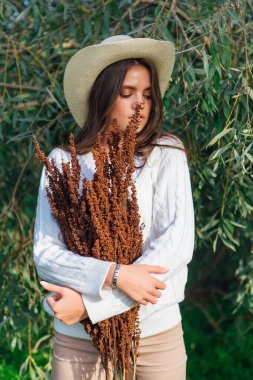 Beyaz kazak, kot pantolon ve kovboy hasır şapkası giymiş genç, güzel esmer kadın elinde kuru yapraklarla ağaç brunch 'larına yakın duruyor.