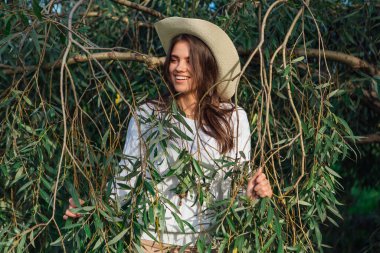 Beyaz kazak, kot pantolon ve kovboy hasır şapkası giymiş genç, güzel esmer kadın yeşil yapraklı ağaç brunch 'larına yakın duruyor.