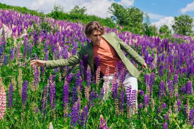 Yeşil ceketli uzun boylu yakışıklı bir adam lupin çiçeği tarlasında dans ediyor, doğanın güzelliğini emrediyor. Mor ve pembe lupinlerle çevrili bir adam..