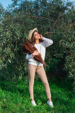 Beyaz kazak, kot pantolon ve kovboy hasır şapkası giymiş genç, güzel esmer kadın elinde kuru yapraklarla ağaç brunch 'larına yakın duruyor.