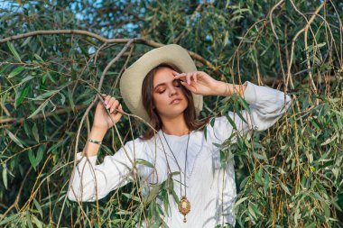 Beyaz kazak, kot pantolon ve kovboy hasır şapkası giymiş genç, güzel esmer kadın yeşil yapraklı ağaç brunch 'larına yakın duruyor.