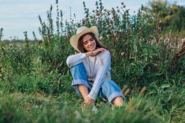 Beyaz kazak, kot pantolon ve kovboy hasır şapkası giyen genç, güzel esmer kadın yeşil çimlerin üzerinde oturuyor, gün batımında gülümsüyor ve gülüyor.