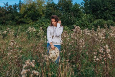 Beyaz kazak, kot pantolon ve kovboy hasır şapkası giymiş genç, güzel esmer kadın kuru kahverengi çimlerin üzerinde duruyor, gün batımında gülümsüyor ve gülüyor.