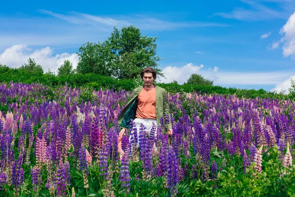 Tall handsome man in a green jacket standing on lupine flowers field with pink cloth, enjoing the beauty of nature. Man surrounded by purple and pink lupines.
