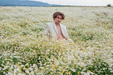 Beyaz takım elbiseli uzun boylu yakışıklı adam papatya çiçeği tarlasında beyaz bir sandalyede oturuyor.