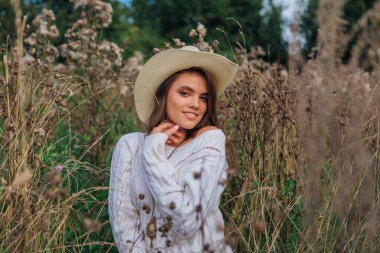 Beyaz kazak, kot pantolon ve kovboy hasır şapkası giyen genç, güzel esmer kadın kuru kahverengi çimlerin üzerinde oturuyor, gün batımında gülümsüyor ve gülüyor.