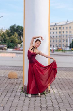 Kırmızı bale elbiseli kadın balerin eski sütunların yanında sivri ayakkabıyla dans ediyor. Balerin güzel bir bale pozisyonunda duruyor.