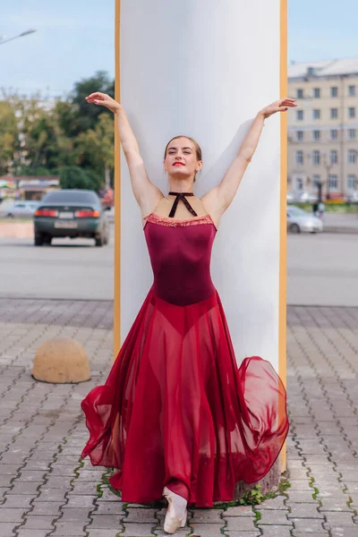 Kırmızı bale elbiseli kadın balerin eski sütunların yanında sivri ayakkabıyla dans ediyor. Balerin güzel bir bale pozisyonunda duruyor.