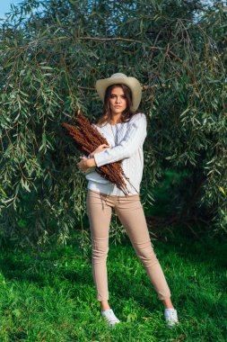 Beyaz kazak, kot pantolon ve kovboy hasır şapkası giymiş genç, güzel esmer kadın elinde kuru yapraklarla ağaç brunch 'larına yakın duruyor.