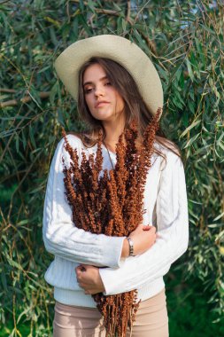 Beyaz kazak, kot pantolon ve kovboy hasır şapkası giymiş genç, güzel esmer kadın elinde kuru yapraklarla ağaç brunch 'larına yakın duruyor.