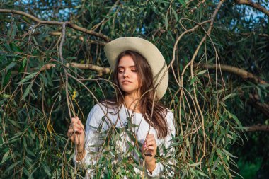 Beyaz kazak, kot pantolon ve kovboy hasır şapkası giymiş genç, güzel esmer kadın yeşil yapraklı ağaç brunch 'larına yakın duruyor.