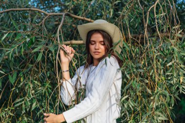 Beyaz kazak, kot pantolon ve kovboy hasır şapkası giymiş genç, güzel esmer kadın yeşil yapraklı ağaç brunch 'larına yakın duruyor.