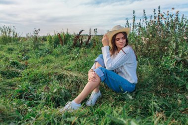 Beyaz kazak, kot pantolon ve kovboy hasır şapkası giyen genç, güzel esmer kadın yeşil çimlerin üzerinde oturuyor, gün batımında gülümsüyor ve gülüyor.