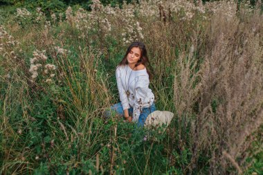 Beyaz kazak, kot pantolon ve kovboy hasır şapkası giyen genç, güzel esmer kadın kuru kahverengi çimlerin üzerinde oturuyor, gün batımında gülümsüyor ve gülüyor.