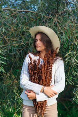 Beyaz kazak, kot pantolon ve kovboy hasır şapkası giymiş genç, güzel esmer kadın elinde kuru yapraklarla ağaç brunch 'larına yakın duruyor.