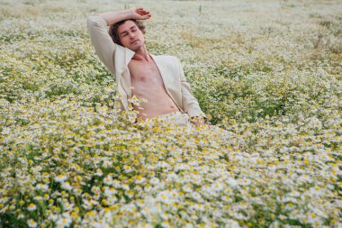 Beyaz takım elbiseli uzun boylu yakışıklı adam papatya çiçeği tarlasında beyaz bir sandalyede oturuyor.