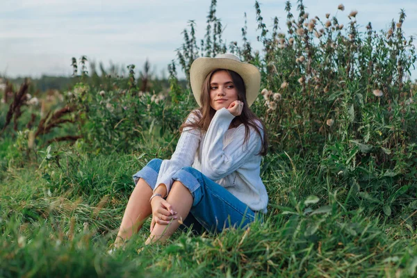 Beyaz kazak, kot pantolon ve kovboy hasır şapkası giyen genç, güzel esmer kadın yeşil çimlerin üzerinde oturuyor, gün batımında gülümsüyor ve gülüyor.