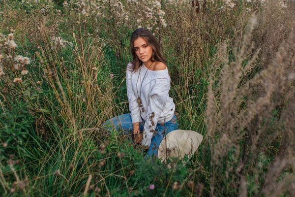 Beyaz kazak, kot pantolon ve kovboy hasır şapkası giyen genç, güzel esmer kadın kuru kahverengi çimlerin üzerinde oturuyor, gün batımında gülümsüyor ve gülüyor.