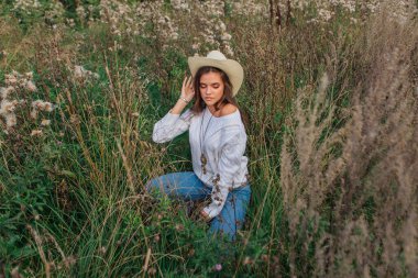 Beyaz kazak, kot pantolon ve kovboy hasır şapkası giyen genç, güzel esmer kadın kuru kahverengi çimlerin üzerinde oturuyor, gün batımında gülümsüyor ve gülüyor.