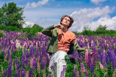 Yeşil ceketli uzun boylu yakışıklı bir adam lupin çiçeği tarlasında duruyor cebinde lupin çiçekleri, doğanın güzelliğini emrediyor. Mor ve pembe lupinlerle çevrili bir adam..