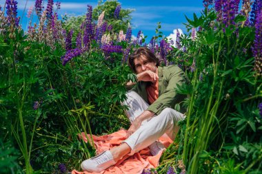 Yeşil ceketli uzun boylu yakışıklı bir adam lupin çiçeklerinin arasında oturmuş, doğanın güzelliğini emrediyor. Mor ve pembe lupinlerle çevrili bir adam..