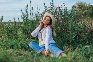 Beyaz kazak, kot pantolon ve kovboy hasır şapkası giyen genç, güzel esmer kadın yeşil çimlerin üzerinde oturuyor, gün batımında gülümsüyor ve gülüyor.