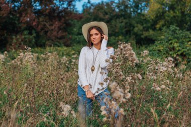 Beyaz kazak, kot pantolon ve kovboy hasır şapkası giymiş genç, güzel esmer kadın kuru kahverengi çimlerin üzerinde duruyor, gün batımında gülümsüyor ve gülüyor.