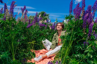 Yeşil ceketli uzun boylu yakışıklı bir adam lupin çiçeklerinin arasında oturmuş, doğanın güzelliğini emrediyor. Mor ve pembe lupinlerle çevrili bir adam..