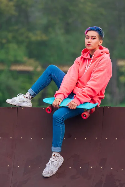 Jong meisje met kort veelkleurig haar zit met skateboard in skatepark — Stockfoto