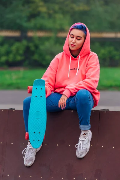 Młoda dziewczyna z krótkimi, wielobarwnymi włosami siedzi z deskorolką w skateparku — Zdjęcie stockowe