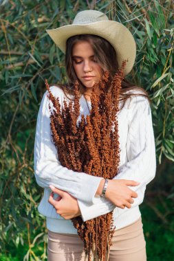 Beyaz kazak, kot pantolon ve kovboy hasır şapkası giymiş genç, güzel esmer kadın elinde kuru yapraklarla ağaç brunch 'larına yakın duruyor.