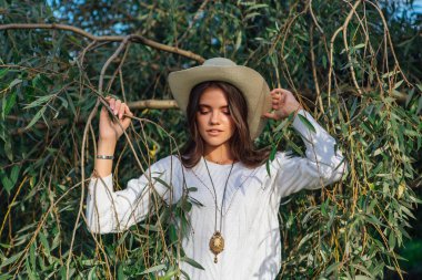 Beyaz kazak, kot pantolon ve kovboy hasır şapkası giymiş genç, güzel esmer kadın yeşil yapraklı ağaç brunch 'larına yakın duruyor.