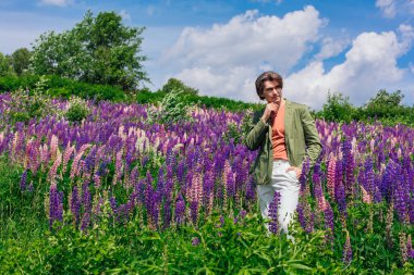 Yeşil ceketli uzun boylu yakışıklı bir adam lupin çiçeği tarlasında duruyor, doğanın güzelliğini emrediyor. Mor ve pembe lupinlerle çevrili bir adam..