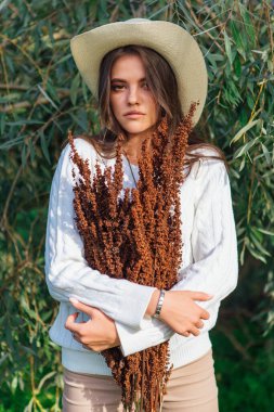 Beyaz kazak, kot pantolon ve kovboy hasır şapkası giymiş genç, güzel esmer kadın elinde kuru yapraklarla ağaç brunch 'larına yakın duruyor.