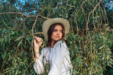 Beyaz kazak, kot pantolon ve kovboy hasır şapkası giymiş genç, güzel esmer kadın yeşil yapraklı ağaç brunch 'larına yakın duruyor.
