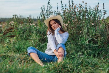 Beyaz kazak, kot pantolon ve kovboy hasır şapkası giyen genç, güzel esmer kadın yeşil çimlerin üzerinde oturuyor, gün batımında gülümsüyor ve gülüyor.