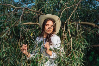 Beyaz kazak, kot pantolon ve kovboy hasır şapkası giymiş genç, güzel esmer kadın yeşil yapraklı ağaç brunch 'larına yakın duruyor.